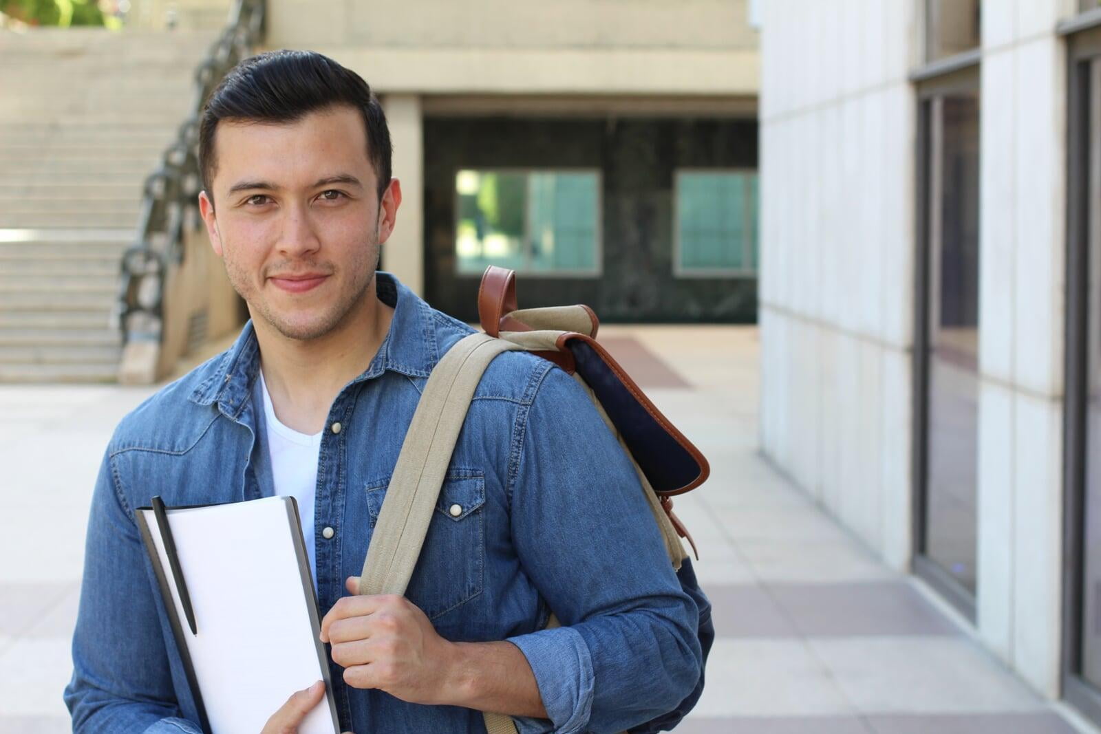 Young adult aged student with backpack and notebook standing outside. 